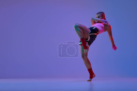 Foto de Foto dinámica de la adolescente atlética, luchadora mma en entrenamiento de ropa deportiva sobre fondo púrpura en luces de neón. Concepto de artes marciales mixtas, deporte, hobby, competición, atletismo, fuerza, ad - Imagen libre de derechos