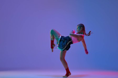 Foto de Foto dinámica de la adolescente deportiva, luchadora mma en entrenamiento de ropa deportiva contra fondo de estudio púrpura en luces de neón. Concepto de artes marciales mixtas, deporte, competición, atletismo, fuerza, ad - Imagen libre de derechos