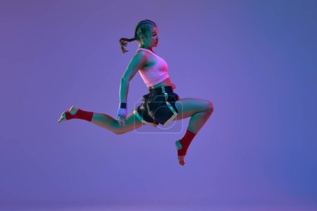 Photo for Kick in a jump. Teen girl, mma athlete in motion, training against purple studio background in neon lights. Concept of mixed martial arts, sport, hobby, competition, athleticism, strength, ad - Royalty Free Image
