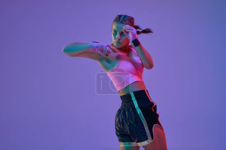 Photo for Defendeign and hitting. Teen girl, MMA fighter in motion, training, boxing against purple studio background in neon lights. Concept of mixed martial arts, sport, hobby, competition, strength, ad - Royalty Free Image