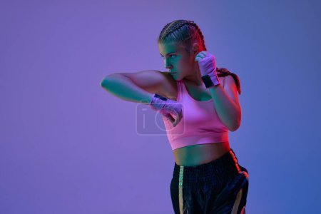 Foto de Chica atlética adolescente, luchador MMA en movimiento, entrenamiento patadas contra fondo estudio púrpura en luces de neón. Concepto de artes marciales mixtas, deporte, hobby, competición, atletismo, fuerza, ad - Imagen libre de derechos