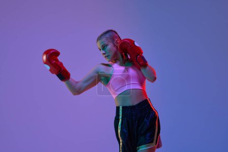 Photo for Sportive teen girl, MMA athlete in uniform and boxing gloves, training against purple background in neon lights. Concept of mixed martial arts, sport, hobby, competition, athleticism, strength, ad - Royalty Free Image