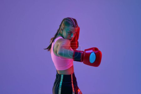 Foto de Chica adolescente deportiva, atleta MMA en uniforme y guantes de boxeo, entrenamiento sobre fondo púrpura en luces de neón. Concepto de artes marciales mixtas, deporte, hobby, competición, atletismo, fuerza, ad - Imagen libre de derechos