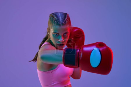 Photo for Competitive teen girl, MMA fighter in motion, kicking in gloves against purple studio background in neon lights. Concept of mixed martial arts, sport, hobby, competition, athleticism, strength, ad - Royalty Free Image