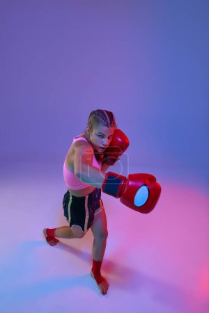 Foto de Adolescente competitiva, luchadora MMA en movimiento, pateando guantes contra fondo púrpura estudio en luces de neón. Concepto de artes marciales mixtas, deporte, hobby, competición, atletismo, fuerza, ad - Imagen libre de derechos