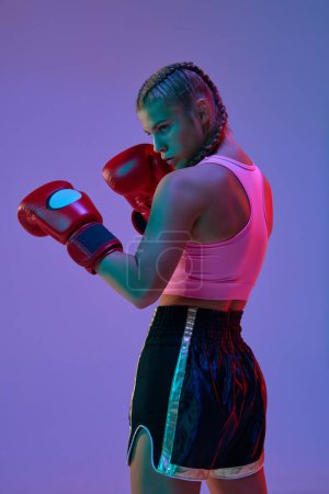Photo for Serious teen girl, MMA fighter in sportswear and boxing gloves, standing in position, training against purple background in neon lights. Concept of mixed martial arts, sport, hobby, competition, ad - Royalty Free Image