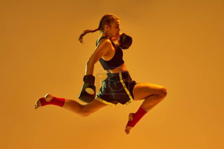 Photo for Kick in a jump. Teen girl, mma athlete in motion, training against orange studio background in neon lights. Concept of mixed martial arts, sport, hobby, competition, athleticism, strength, ad - Royalty Free Image