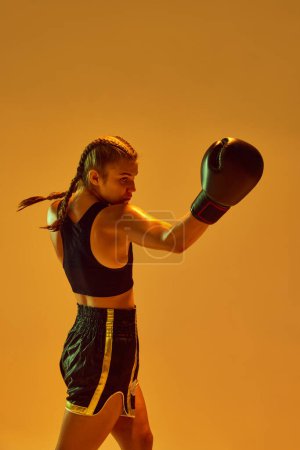 Photo for Sportive teen girl, MMA athlete in uniform and boxing gloves, training against orange background in neon lights. Concept of mixed martial arts, sport, hobby, competition, athleticism, strength, ad - Royalty Free Image