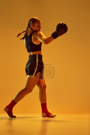 Téléchargez les photos : Athlétique, jeune fille sérieuse et concentrée, entraînement de combat MMA, lutte contre fond de studio orange aux néons. Concept d'arts martiaux mixtes, sport, passe-temps, compétition, force, publicité - en image libre de droit