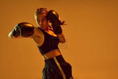Photo for Sportive teen girl, MMA athlete in uniform and boxing gloves, training against orange studio background in neon lights. Concept of mixed martial arts, sport, hobby, competition, athleticism, strength - Royalty Free Image