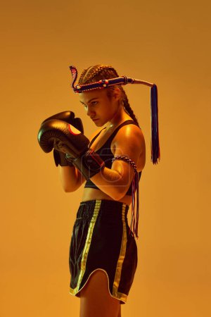Photo for Defense position. Teen girl, concentrated MMA athlete in boxing gloves training against orange studio background in neon lights. Concept of mixed martial arts, sport, hobby, competition, strength, ad - Royalty Free Image