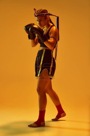 Téléchargez les photos : Adolescente, combattante MMA en uniforme et gants de boxe, debout en position, s'entraînant sur fond de studio orange aux néons. Concept d'arts martiaux mixtes, sport, compétition, force, publicité - en image libre de droit