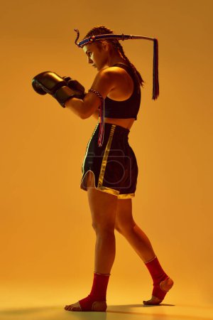 Photo for Teen girl, MMA fighter in uniform and boxing gloves, standing in position, training against orange studio background in neon lights. Concept of mixed martial arts, sport, competition, strength, ad - Royalty Free Image