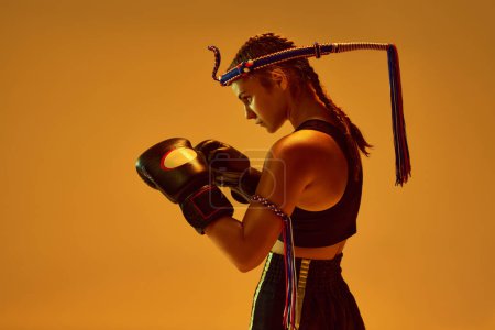 Photo for Defense position. Teen girl, concentrated MMA athlete in boxing gloves training against orange studio background in neon lights. Concept of mixed martial arts, sport, hobby, competition, strength, ad - Royalty Free Image