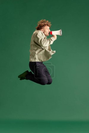 Photo for Mature man in casual clothes emotionally shouting in megaphone and jumping against green studio background. News, sales. Concept of human emotions, facial expression, lifestyle, fashion, ad - Royalty Free Image