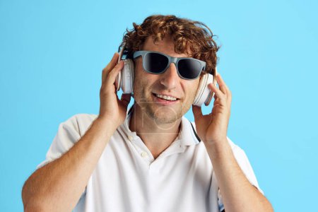 Téléchargez les photos : Homme mûr en t-shirt blanc décontracté et lunettes de soleil écoutant de la musique dans un casque sur fond de studio bleu. Le plaisir. Concept d'émotions humaines, expression faciale, mode de vie - en image libre de droit