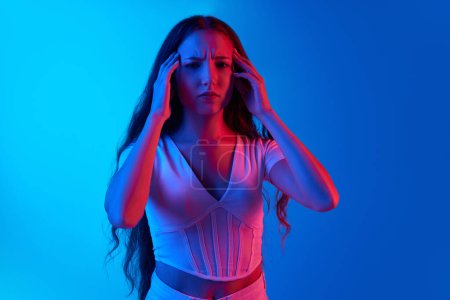 Photo for Young woman holding hands on head, suffering from headache and stress against blue studio background in neon light. Concept of human emotions, facial expression, beauty, lifestyle, youth, ad - Royalty Free Image