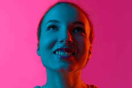 Téléchargez les photos : Gros plan portrait de jeune fille souriante regardant vers le haut sur fond de studio rose sous la lumière du néon. Concept d'émotions humaines, mode, beauté, mode de vie, jeunesse, publicité - en image libre de droit