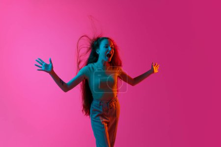 Foto de Chica joven de pie con el viento soplando en el pelo, gritando, posando emocionalmente sobre fondo de estudio rosa en luz de neón. Concepto de emociones humanas, moda, belleza, estilo de vida, juventud, anuncio - Imagen libre de derechos