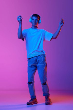 Foto de Longitud completa de chico joven escuchando música en auriculares y bailando sobre el color rosa degradado fondo púrpura en luz de neón. Concepto de emociones humanas, juventud, estilo de vida, moda, expresiones faciales, anuncio - Imagen libre de derechos