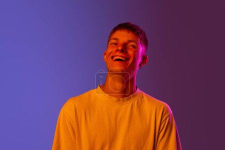 Foto de Retrato de un joven riéndose alegremente contra el degradado fondo púrpura en luz de neón. Felicidad y alegría. Concepto de emociones humanas, juventud, estilo de vida, moda, expresiones faciales, anuncio - Imagen libre de derechos