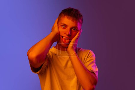 Photo for Young emotional guy posing with funny, meme, grimacing face against gradient purple background in neon light. Concept of human emotions, youth, lifestyle, fashion, facial expressions, ad - Royalty Free Image