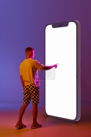 Foto de Hombre joven desplazándose pantalla gigante 3d del teléfono móvil contra el gradiente de fondo púrpura en luz de neón. Concepto de emociones humanas, juventud, estilo de vida, moda, compras en línea, ventas, anuncios - Imagen libre de derechos