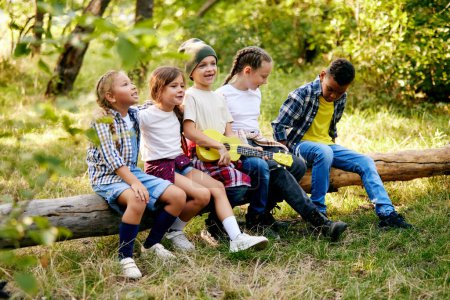 Téléchargez les photos : Enfants joyeux et ludiques assis sur le tronc dans la forêt par une journée ensoleillée. Randonnées pédestres, promenades, week-ends actifs. Concept d'activité de loisirs, enfance, été, amitié, mode de vie actif, plaisir, nature - en image libre de droit