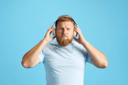 Téléchargez les photos : Homme rousse barbu écoutant de la musique dans un casque sur fond de studio bleu. Concept d'émotions humaines, mode de vie, expression faciale, annonce. Espace de copie pour la publicité - en image libre de droit