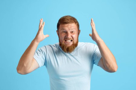 Photo for Bearded redhead man emotionally shouting, expressing anger and irritation against blue studio background. Concept of human emotions, lifestyle, facial expression, ad. Copy space for ad - Royalty Free Image