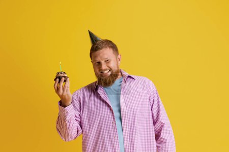 Photo for Happy, smiling, bearded man standing with cake, celebrating birthday against yellow studio background. Concept of human emotions, lifestyle, party, celebration, sales, fun, ad. Copy space for ad - Royalty Free Image