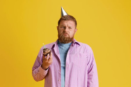Photo for Sad bearded man standing with cake, celebrating birthday against yellow studio background. Aging. Concept of human emotions, lifestyle, party, celebration, sales, fun, ad. Copy space for ad - Royalty Free Image