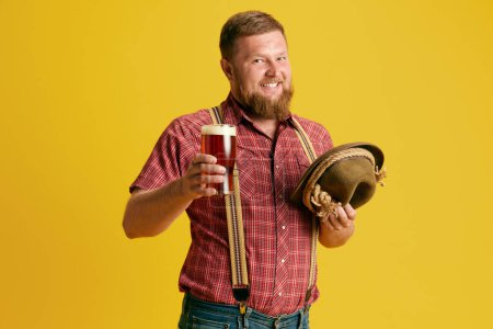 Photo for Smiling, bearded man in checkered shirt standing with beer glass against yellow studio background. Concept of human emotions, lifestyle, festival, party, oktoberfest, ad. Copy space for ad - Royalty Free Image