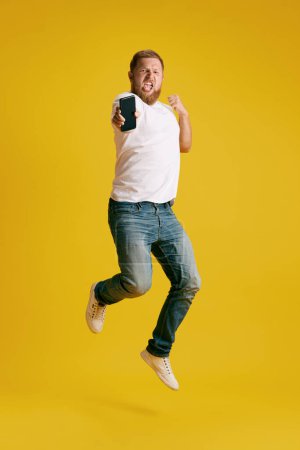 Photo for Man emotionally jumping with mobile phone against yellow studio background. Success, winning. Concept of emotions, lifestyle, business, freelance, job fair, betting, sales, ad. Copy space for ad - Royalty Free Image