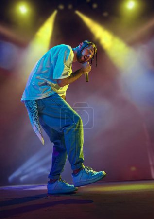 Photo for Young artistic man, musician making live concert, singing, dancing on stage over colorful neon illumination. Night club. Music, performance, art, talent, nightlife, joy, party and lifestyle concept - Royalty Free Image