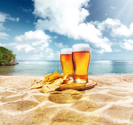 Foto de Dos vasos de cerveza espumosa, fría, cerveza de cerveza con papas fritas aperitivos en la arena contra el océano y el fondo azul del cielo. Concepto de cerveza, cervecería, vacaciones y vacaciones, tradiciones, festival, bebida alcohólica, anuncio - Imagen libre de derechos