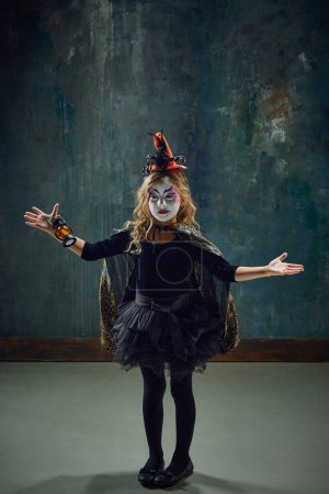 Téléchargez les photos : Petite fille, enfant avec maquillage de sorcière coloré et costume debout sur fond vintage sombre. Mystère. Concept d'Halloween, enfance, célébration, fête, vacances, créativité, annonce - en image libre de droit