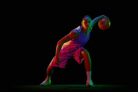 Photo for Concentrated young guy in sportswear playing basketball, training dribbling ball exercises against black background in neon light. Concept of professional sport, competition, hobby, game, competition - Royalty Free Image