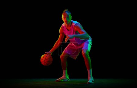 Photo for Dynamic image of teen boy in uniform, playing basketball against black studio background in neon light. Dribbling ball. Concept of professional sport, competition, hobby, game, competition - Royalty Free Image