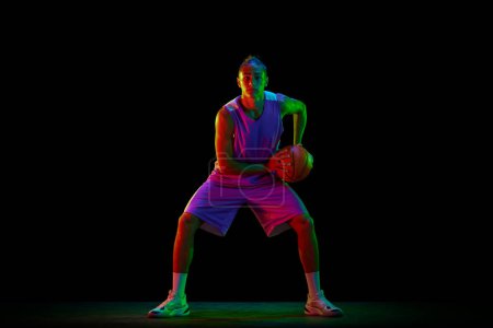 Photo for Young, active, motivated guy, basketball player in uniform standing in position with ball against black background in neon light. Concept of professional sport, competition, hobby, game, competition - Royalty Free Image
