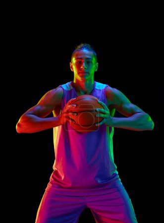 Photo for Handsome teen boy, basketball player in uniform standing with ball and smiling against black studio background in neon light. Concept of professional sport, competition, hobby, game, competition - Royalty Free Image