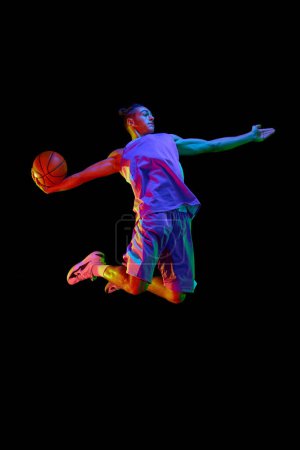 Foto de Imagen dinámica del joven, jugador de baloncesto en uniforme en movimiento, saltando con pelota sobre fondo negro estudio en luz de neón. Concepto de deporte profesional, competición, hobby, juego, competición - Imagen libre de derechos