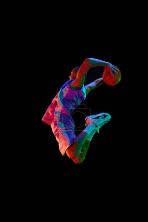 Photo for Athletic young guy, basketball player in motion during match, jumping with ball against black studio background in neon light. Concept of professional sport, competition, hobby, game, competition - Royalty Free Image