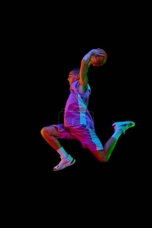 Photo for Scoring winning goal. Young man in uniform playing basketball, jumping with ball against black studio background in neon light. Concept of professional sport, competition, hobby, game, competition - Royalty Free Image