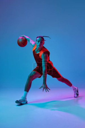 Téléchargez les photos : Un jeune homme concentré en vêtements de sport jouant au basket, s'entraînant à dribbler des exercices de balle sur fond de studio bleu sous la lumière du néon. Concept de sport professionnel, compétition, passe-temps, jeu, compétition - en image libre de droit