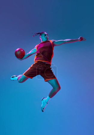 Téléchargez les photos : Entraînement sportif et compétitif de garçons adolescents, jouant au basket-ball sur fond de studio bleu sous la lumière du néon. Concept de sport professionnel, compétition, passe-temps, jeu, compétition - en image libre de droit