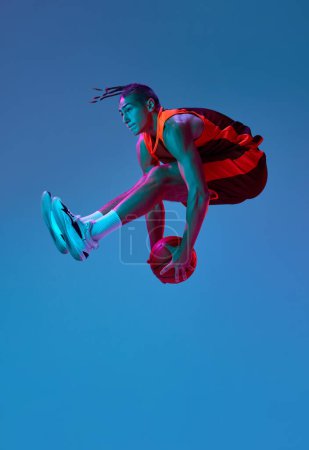 Photo for Sportive young guy, basketball player in motion during match, jumping with ball against blue studio background in neon light. Concept of professional sport, competition, hobby, game, competition - Royalty Free Image