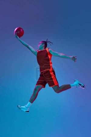 Téléchargez les photos : Pleine longueur de jeune homme compétitif en mouvement pendant le match de basket-ball, sautant avec une balle sur fond bleu studio sous la lumière du néon. Concept de sport professionnel, compétition, passe-temps, jeu, compétition - en image libre de droit