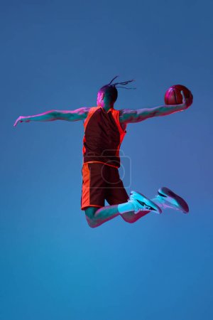 Photo for Scoring winning goal. Young man in uniform playing basketball, jumping with ball against blue studio background in neon light. Concept of professional sport, competition, hobby, game, competition - Royalty Free Image