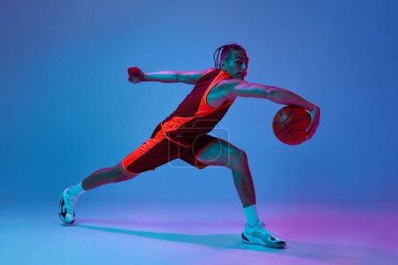 Téléchargez les photos : Un jeune homme concentré en vêtements de sport jouant au basket, s'entraînant à dribbler des exercices de balle sur fond de studio bleu sous la lumière du néon. Concept de sport professionnel, compétition, passe-temps, jeu, compétition - en image libre de droit